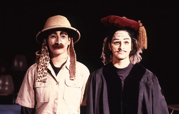 Two people in theatrical costumes stand together. The person on the left wears a pith helmet, leopard print accessory, and mustache. The person on the right wears a red beret with a tassel, black robe, and painted face.