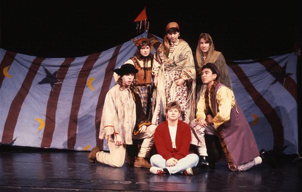 A group of six people, dressed in various costumes, pose on a stage. A backdrop painted with moons and stars is behind them. One person sits cross-legged in front, while others stand or kneel around. They appear to be engaged in a performance.