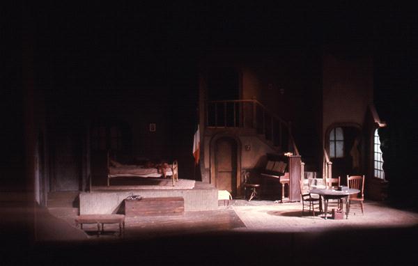 Dimly lit stage set depicting an interior room with a bed, a piano, a table with chairs, and a small staircase leading to an upper level. The muted colors and shadows create a somber atmosphere.