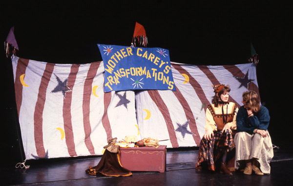 Two actors in costume sit on stage in front of a striped and starry backdrop with the banner "Mother Carey's Transformations." Props are placed on the stage, including a chest and draped fabric near the performers.