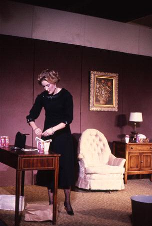 A person in a black dress is standing at a table with food containers. The scene is set in a room with a beige armchair, a wooden side table, a lamp, and a framed painting on the wall.