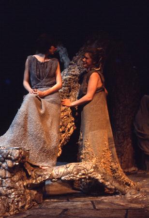 Two women in textured, earth-toned costumes stand on a stage with a rugged, organic set design. One woman sits on a branch-like structure, while the other stands and leans in, engaging in conversation. The lighting is dim and dramatic.