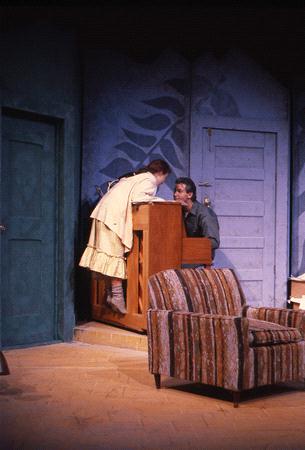 Two performers on stage, with one playing an upright piano while the other leans closely over it. They are in a room with a green door and a patterned blue wall. A striped armchair is in the foreground.