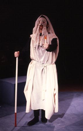 Person dressed in ancient Greek-style costume, wearing a white robe and mask with long white hair and beard. They hold a staff and stand in a dimly lit setting with dark background.