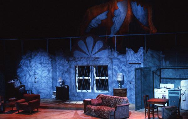 A theater stage set depicts a vintage living room with a floral couch, red armchair, and side table with a lamp. A kitchen area with a fridge and stove is on the right. Blue lighting casts dramatic shadows, creating a mysterious atmosphere.