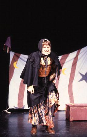 A person dressed as a witch or fortune teller stands on stage wearing a dark cloak and headscarf. They are in front of a curtain decorated with moons and stars, with a small wooden box beside them.