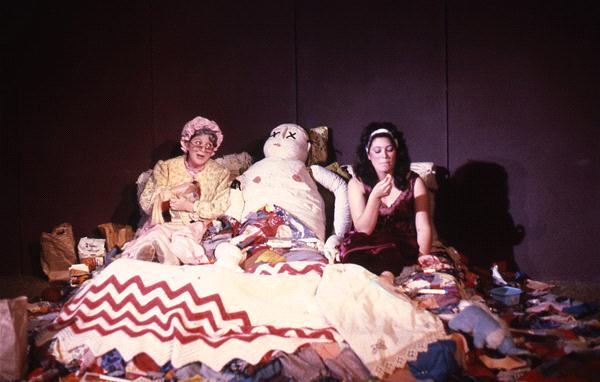 Two people sit on a cluttered bed, surrounded by various items. The person on the left wears a patterned outfit and a hat, while the person on the right is in a dark dress. In between them is a large stuffed figure.