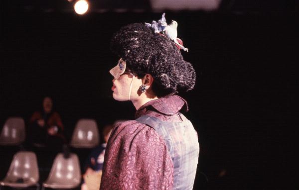 A person in profile wearing a masquerade mask and an elaborate outfit with a textured purple top and vest. The hairstyle includes decorative elements. They are in a room with an audience seated on white chairs in the background.