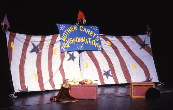 A stage setup with a large, colorful backdrop featuring stars and moons. A blue banner reads "Mother Carey's Transformations." Two boxes and a draped cloth sit on the stage floor.