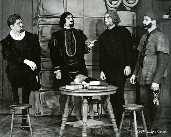 Four men in historical costumes stand and sit around a table with books and a helmet in a room with stone walls. One man points as they engage in conversation. They have various styles of facial hair and wear robes, a tunic, and a hat.