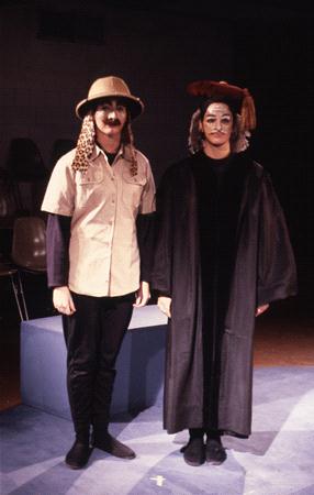 Two people in costume stand on a stage. One wears an explorer outfit with a leopard hat, and the other is dressed in a black robe with a brown hat. Both have painted faces. They are standing on a blue platform with chairs in the background.