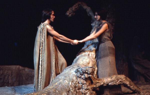 Two people holding hands in a theatrical setting with a large, gnarled tree between them. Both are dressed in earth-toned, flowing costumes. The atmosphere is dark and dramatic, suggesting a scene of ancient or mythical significance.