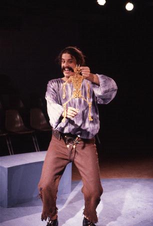 A person on a theatrical stage energetically holding a small skeleton prop, wearing a loose-fitting purple shirt and brown pants. The background is dimly lit with a few empty chairs visible, suggesting a performance setting.