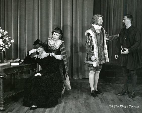 Black and white photo of four people in period costumes performing on stage. One person is seated at a table, another consoles them, and two stand nearby in conversation. A curtain is in the background, and a vase of flowers is on the table.
