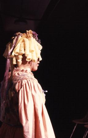 A person wearing a vintage, ruffled dress and a bonnet adorned with ribbons and flowers stands onstage against a dark background, facing sideways. The lighting creates a dramatic contrast, highlighting the details of the outfit.