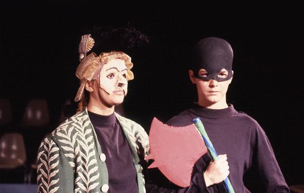 Two individuals in theatrical costumes on stage. One wears a decorative hat and patterned cape, holding a red and blue staff. The other is in black with a matching mask and holding a red axe. Background features empty chairs.