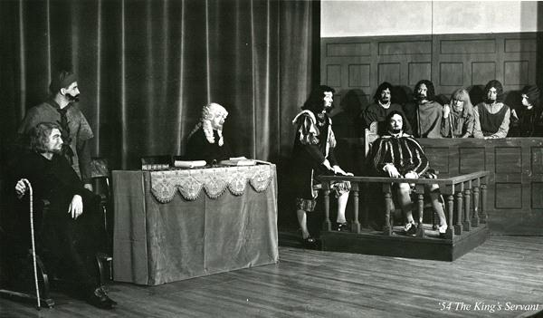 A black-and-white photo of a staged courtroom scene with actors in period costumes. A judge sits at a table, while a man stands in the witness box. Others are seated in the courtroom as observers, with wood paneling and curtains in the background.