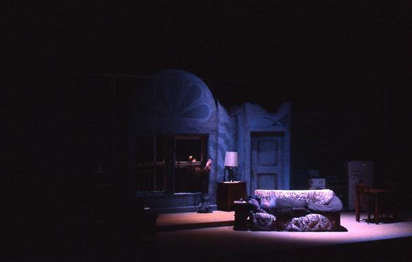 Dimly lit room with a vintage vibe, featuring a couch, a floor lamp beside a door, and a person looking out a window. The setting suggests a dramatic or theatrical atmosphere, with deep shadows and a sense of mystery.