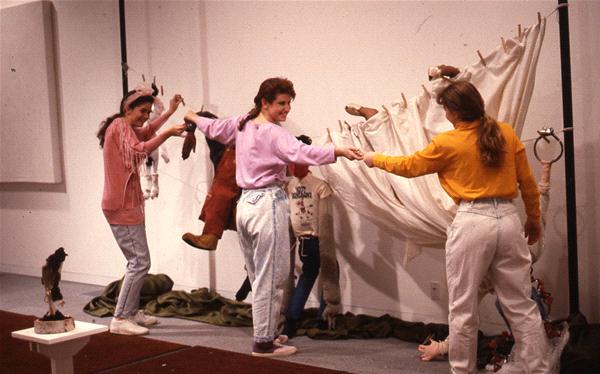 Four people in casual clothing interact with a sculptural installation featuring fabric and objects resembling body parts. The room is plain with white walls and carpeting.