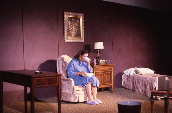 A person in a blue robe sits in an armchair, sipping from a cup. The room has a bed, wooden side table with a lamp, and a framed picture on the wall. A small TV is in the foreground. The setting resembles a cozy, modest living space.