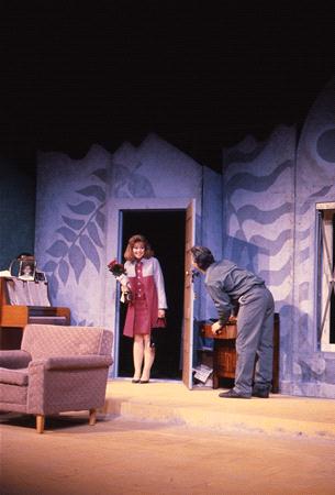 A woman in a red dress stands in a doorway holding flowers, looking at a man in a gray outfit who is crouching by a cabinet. The stage set includes a sofa and a blue wall with abstract designs.