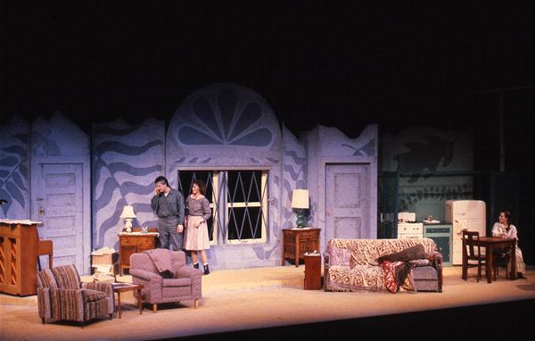 A stage set depicts a mid-20th century living room with two people standing near a window. The room has a sofa, chairs, and tables with lamps. On the right, a person sits at a kitchen table. The backdrop features ocean waves and a fan design.