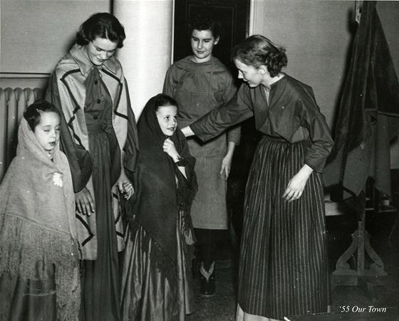 A black and white photo showing five people. Two adults, dressed in skirts and shawls, are with three children. One adult stands beside a child, adjusting her shawl. They appear to be dressed in period costumes. "’55 Our Town" is written in the corner.