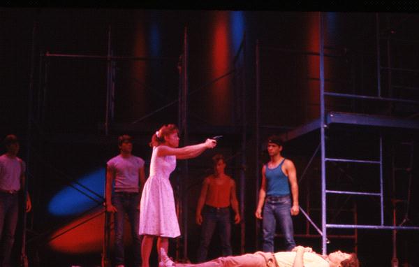 A woman in a white dress points a gun on stage, surrounded by four men. One man lies on the ground. The backdrop features scaffolding and colorful lighting, creating a dramatic atmosphere.