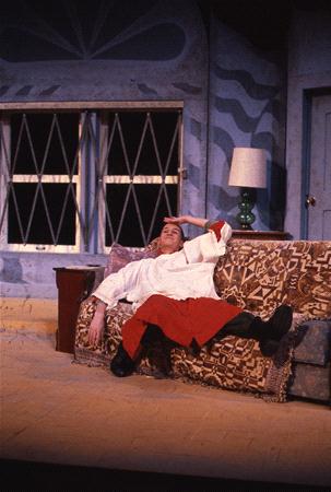 Person in a red and white costume lies dramatically on a patterned couch in a stage set, with one hand on their forehead. The background includes abstract geometric windows and a lamp with a green base.