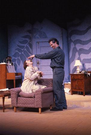 A man stands in front of a woman seated on a couch on a stage. The man reaches towards the woman's head. The setting includes a piano, small table with a lamp, and a painted backdrop, creating a theatrical scene.