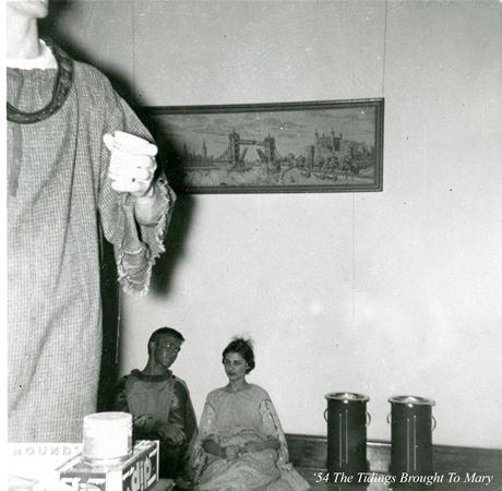 A black and white photo of two people sitting on the floor in medieval-style costumes beneath a framed painting of a bridge. Another person stands nearby, partially out of frame, holding a cup. Two large cylindrical objects are beside the seated pair.