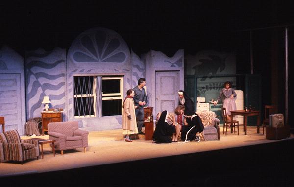 A theater production featuring six actors on stage, with a backdrop of stylized wall designs and furniture including chairs, a table, and a lamp. The set suggests an interior setting, possibly a living room. The actors are engaged in a scene.