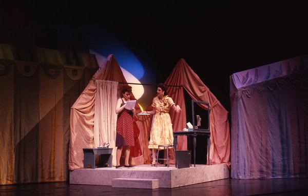 Two actors perform on stage dressed in vintage costumes. One is reading a paper, and the other gestures animatedly. The set resembles a 1950s room with draped curtains and a vanity table, lit with warm, theatrical lighting.