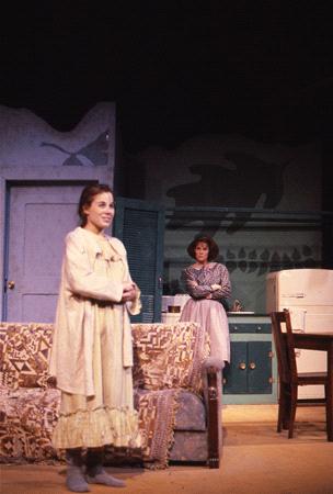 Two actors on a stage set resembling a mid-20th century kitchen. One stands near the fridge in period clothing, while the other, in a nightgown, stands near a sofa. The backdrop features shadows suggesting a domestic setting.