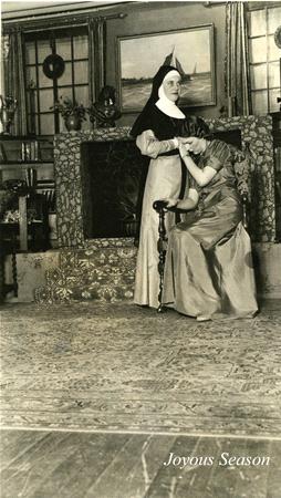 A nun in a traditional habit stands beside a woman in a long dress, who sits with her head resting on the nun's hand. The scene is set in a room with a fireplace and various decorative items. The words "Joyous Season" appear in the corner.
