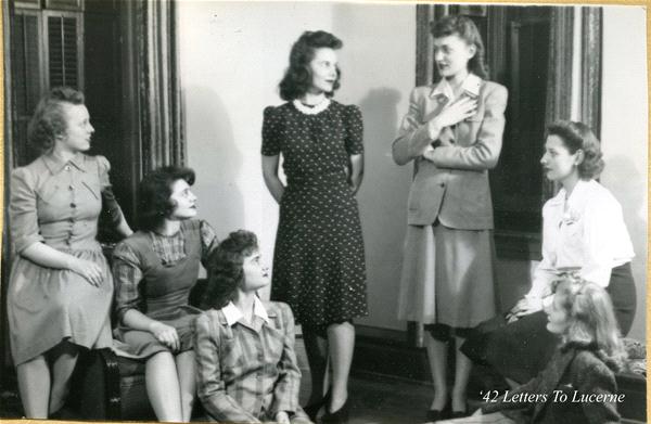 A group of seven women from the 1940s are gathered indoors, engaged in conversation. They wear period dresses and hairstyles, with one standing and gesturing while the others sit or stand around her. Text in the corner reads "'42 Letters to Lucerne.