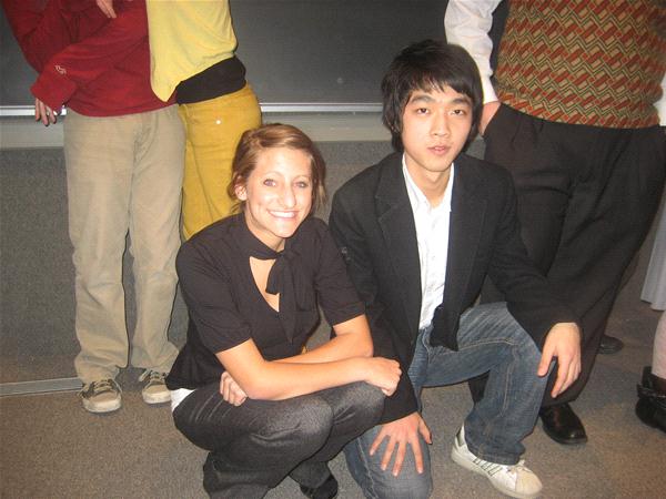 Two people are crouching and smiling indoors. The woman on the left has brown hair and is wearing a black shirt. The man on the right has black hair and is wearing a black jacket and jeans. Other people in the background are partially visible.