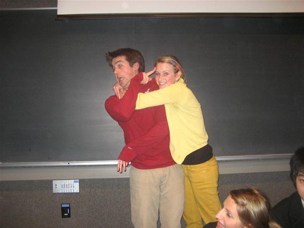 A woman in a yellow sweater playfully pretends to strangle a man in a red sweater. The man has an exaggerated surprised expression. They are in a room with a blackboard in the background. Other people are partially visible around them.