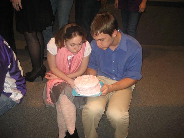 A young woman and man sitting on a carpeted step, both focused on a small pink cake the man holds on his lap. The woman wears a pink scarf and skirt; the man wears a blue shirt and khakis. Other people's legs are visible in the background.