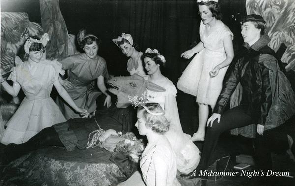 A black and white photo of a theatrical scene from "A Midsummer Night's Dream." Several women dressed as fairies gather around a person lying on the ground. A character in a dark cape stands to the side. The set has a woodland backdrop.