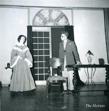 Two people in vintage attire are on stage in a scene from "The Heiress." One person is standing near a chair, wearing a long dress, while the other is in a suit, standing behind a wooden table. A table lamp is visible.