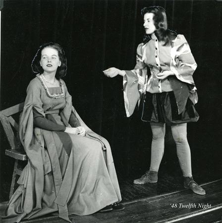 A black-and-white photo of two people performing a scene from "Twelfth Night." One is seated in a long gown, the other stands wearing a tunic and tights, gesturing expressively. They're on a stage with a dark curtain backdrop.