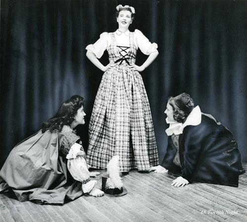 Three actors are performing on stage against a dark curtain. One stands confidently in a plaid dress with hands on hips, while the other two kneel, dressed in period costumes with capes. The scene is theatrical and expressive.