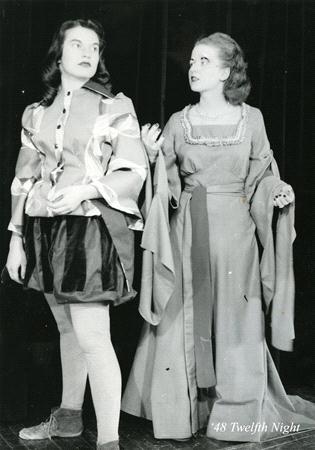 Two actors in theatrical costume perform a scene from "Twelfth Night." One wears a doublet and striped shorts, while the other wears a long gown with wide sleeves. Both look expressive, standing on a dark stage.