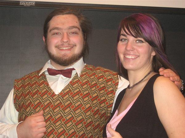 A man with a beard, wearing a patterned vest and bow tie, stands next to a woman with dark hair and purple streaks. They are smiling and standing close together in front of a dark backdrop.