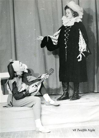 Two actors in period costumes perform on stage. One, seated, wears a jester's outfit and plays a lute. The other, standing, wears a long robe and hat, gesturing with one hand. The scene appears to be from Shakespeare's "Twelfth Night.