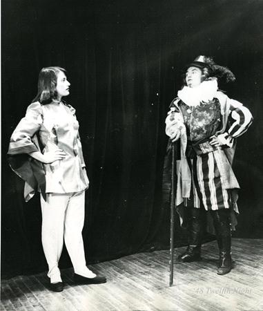 Two actors in period costumes perform on stage. The actor on the left wears a tunic and tights, while the actor on the right wears a hat and ruffled collar, holding a cane. They stand against a dark curtain backdrop.