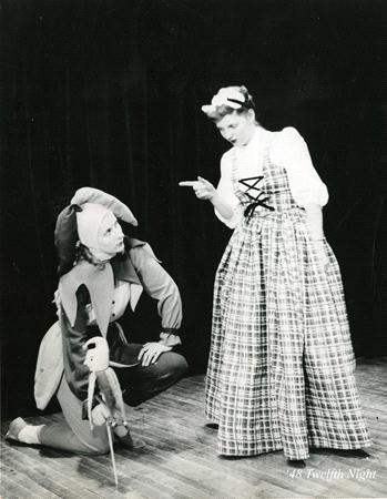 A person in a jester costume kneels on stage, holding a stick with a puppet. Another person wearing a plaid dress and headscarf stands, pointing at the jester. The scene is theatrical with a classic, vintage ambiance. A caption reads "48 Twelfth Night.