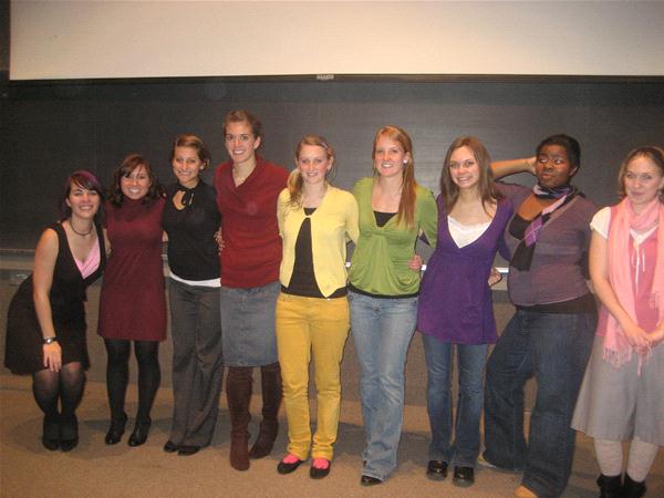 A group of nine people stands in a line in front of a classroom chalkboard, smiling for the camera. They are wearing a variety of outfits, including dresses, skirts, and jeans.