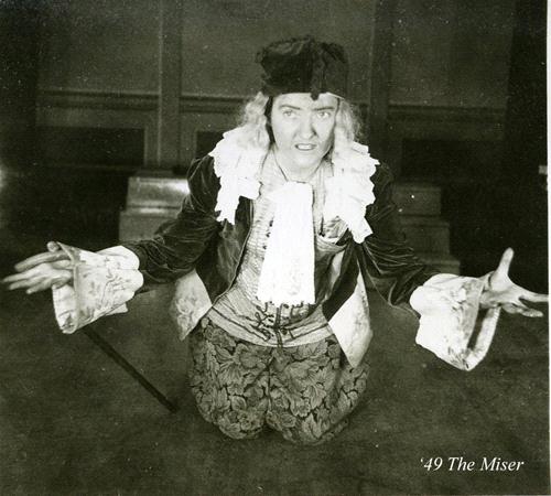 A person in period costume kneels on the ground with arms spread wide. They wear a velvet jacket, lace cuffs, a ruffled shirt, and a hat. The background is a dimly lit room. The text reads '49 The Miser.'.